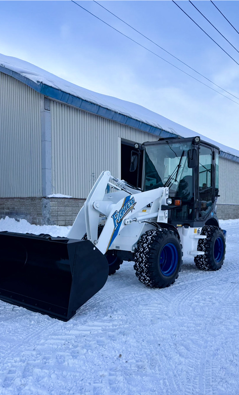 雪の中の工場に停車する除雪用重機：冬のインフラと地域生活を支える活動